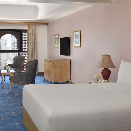 Intercontinental Dar Al Iman Madinah By Ihg Hotel Medina Exterior foto The photo shows a well-furnished hotel room. It features a large bed with a white duvet, two bedside lamps, and decorative paintings on the walls. There is a comfortable seating area with two chairs and a small table. A wooden dresser is positioned a