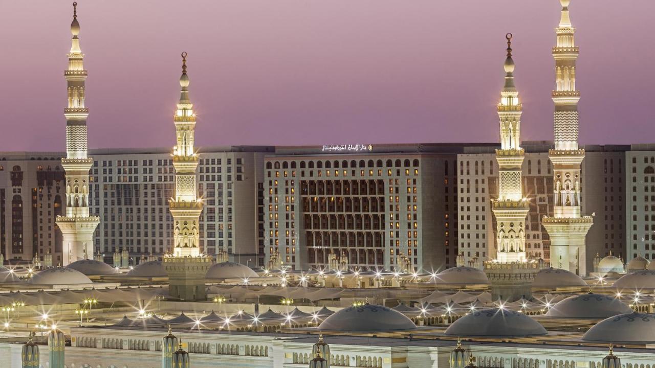 Intercontinental Dar Al Iman Madinah By Ihg Hotel Medina Exterior foto The photo depicts an architectural scene, likely from a mosque complex, featuring prominent minarets that are illuminated. The setting appears to be during twilight, with a soft gradient in the sky. In the background, a large building is visible, pos
