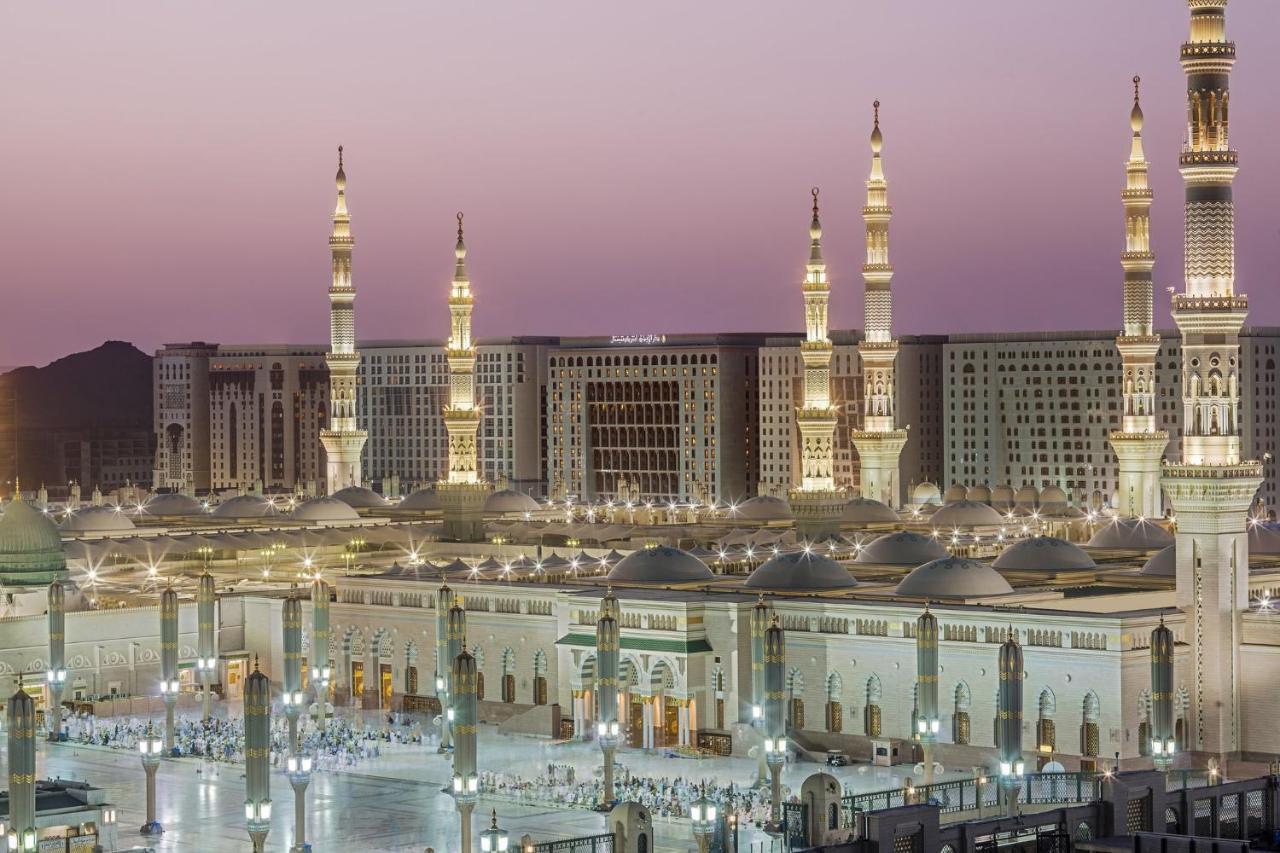 Intercontinental Dar Al Iman Madinah By Ihg Hotel Medina Exterior foto The photo shows a view of an expansive mosque complex, featuring several tall minarets that are illuminated against a twilight sky. The structure appears grand and is likely adorned with intricate architectural details. There are people gathered in t