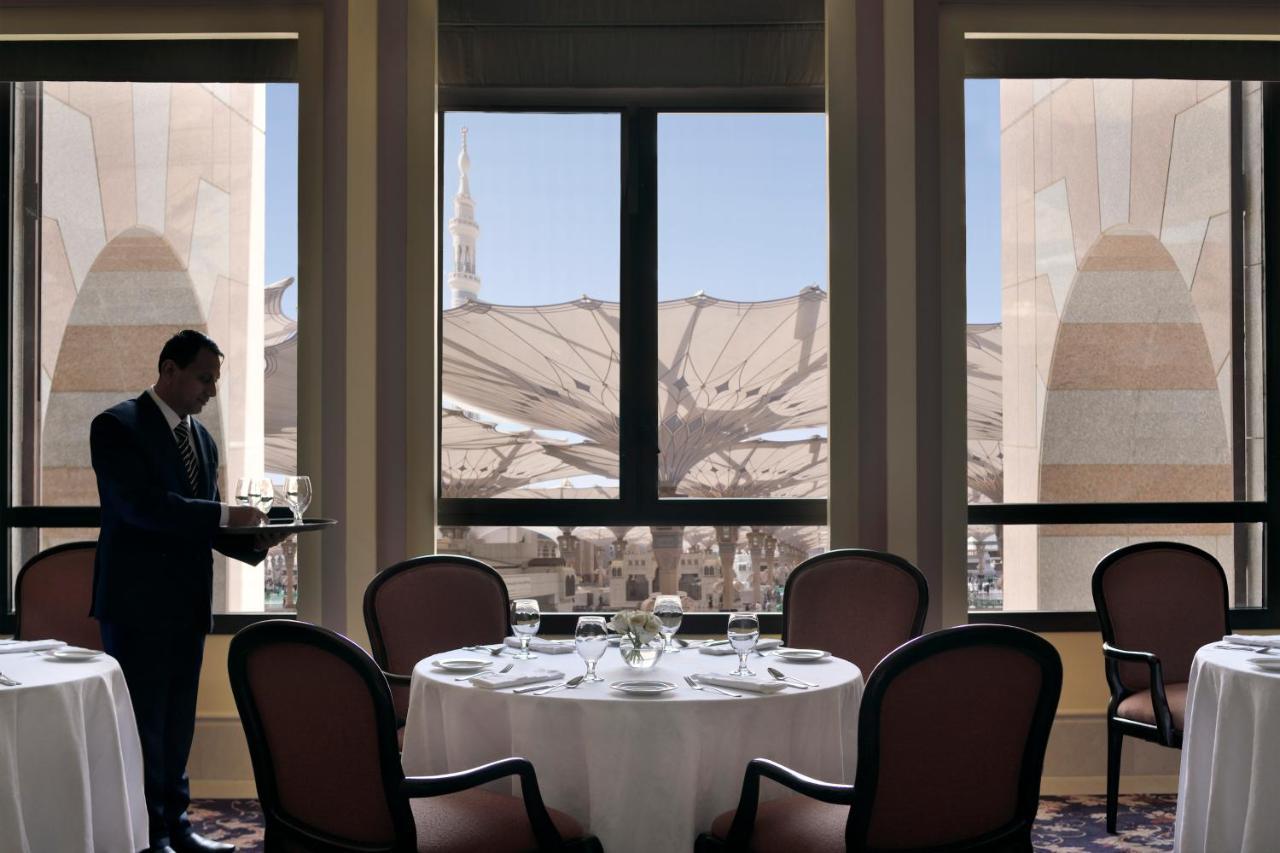 Intercontinental Dar Al Iman Madinah By Ihg Hotel Medina Exterior foto The photo shows a dining area with elegant decor. A waiter is standing to the side, holding a tray. The tables are neatly set with white tablecloths and fine dining arrangements. Large windows provide a view of an outdoor area featuring architectural
