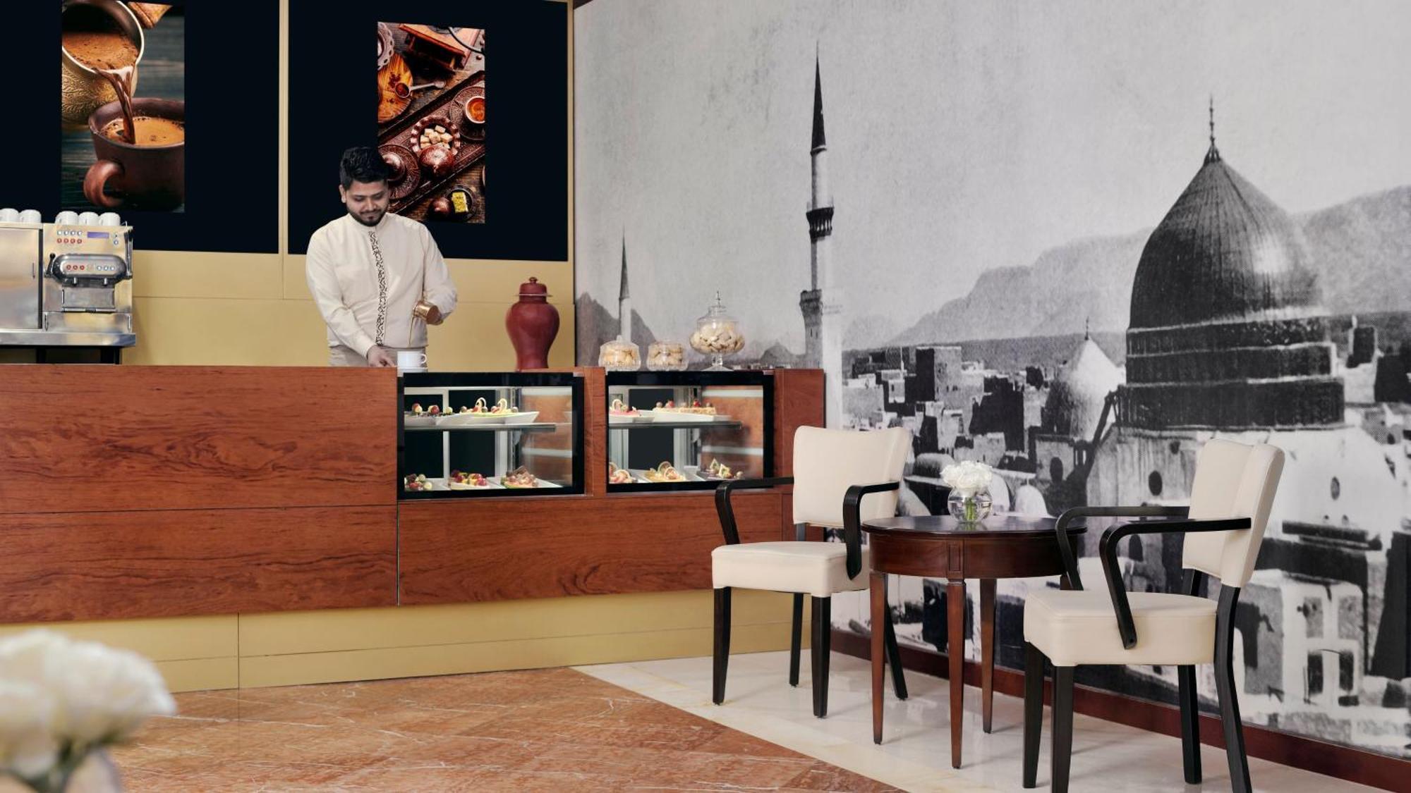 Intercontinental Dar Al Iman Madinah By Ihg Hotel Medina Exterior foto The photo shows an elegant cafe or dessert shop interior. A man in traditional attire is standing behind a wooden counter, where various pastries and sweets are displayed. The walls feature a large black-and-white mural of a cityscape, possibly depic