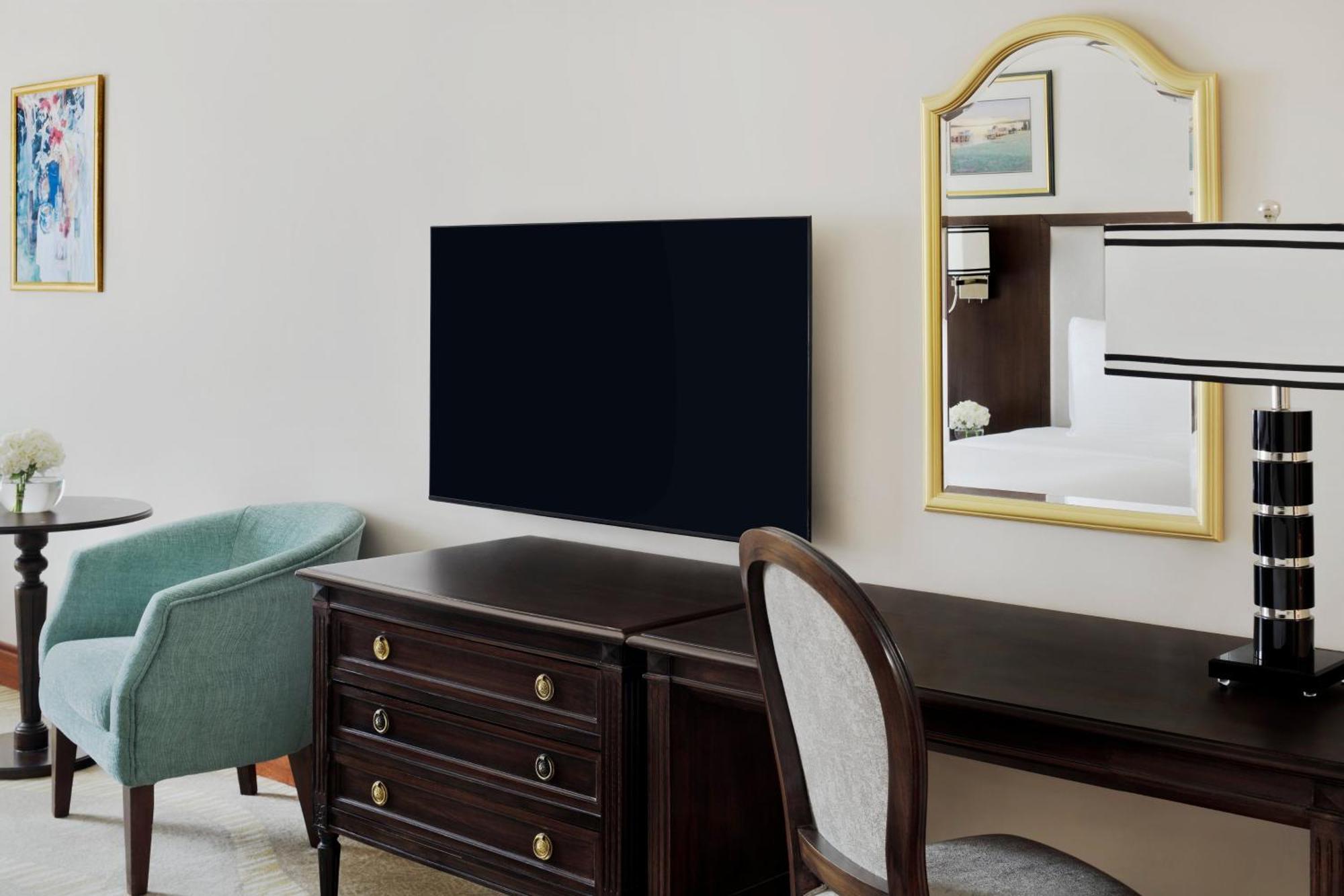 Intercontinental Dar Al Iman Madinah By Ihg Hotel Medina Exterior foto The photo shows a hotel room interior featuring a mounted flat-screen television on the wall. Below the TV, there is a dark wooden dresser with drawers. To the right, there's a wooden desk and a chair with a light-colored seat. A small table with a l