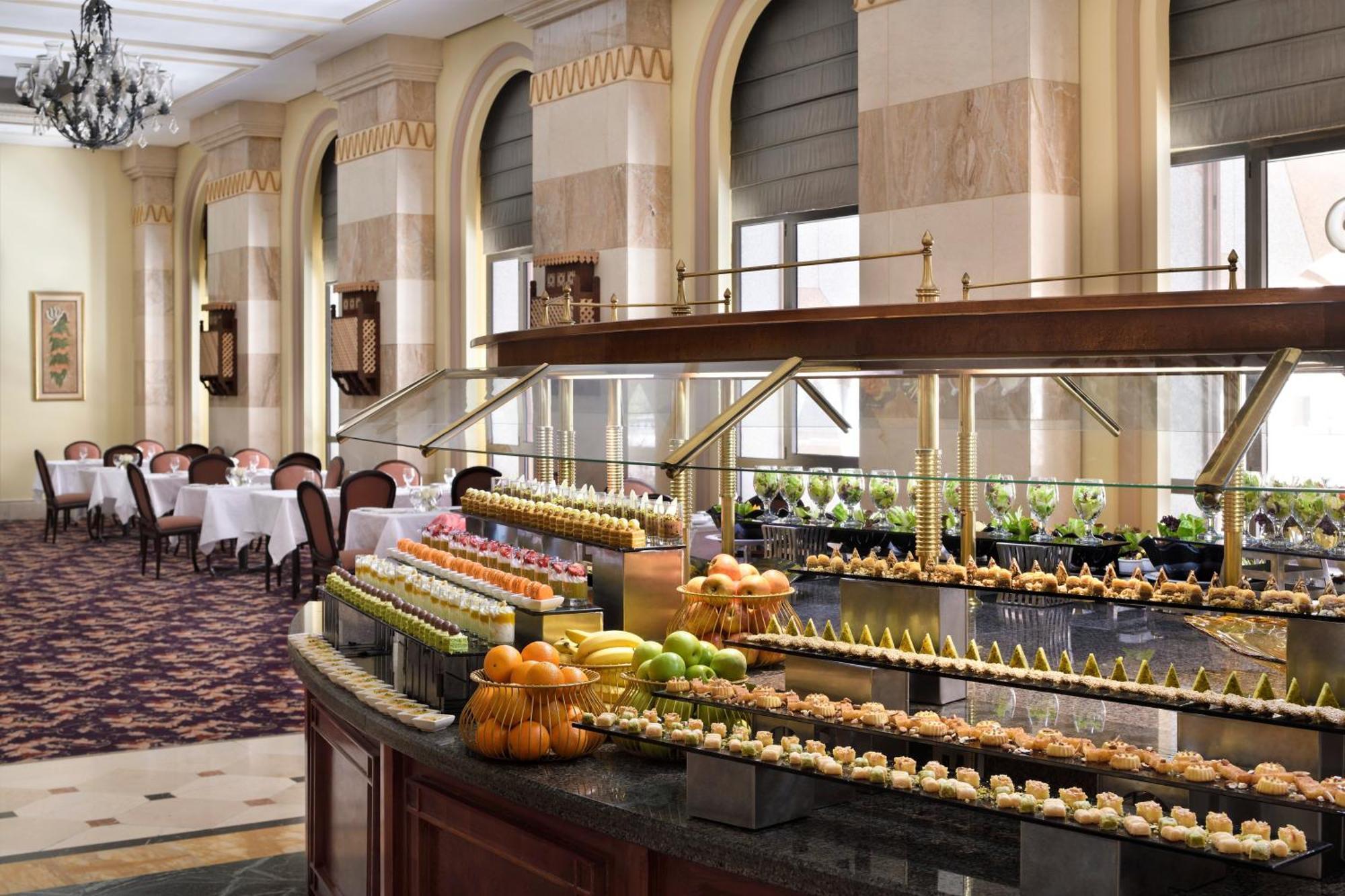 Intercontinental Dar Al Iman Madinah By Ihg Hotel Medina Exterior foto The photo depicts an elegant dining area featuring a buffet-style display. In the foreground, a beautifully arranged buffet counter showcases an array of desserts, including miniature pastries and colorful fruit. The setting has a sophisticated ambia
