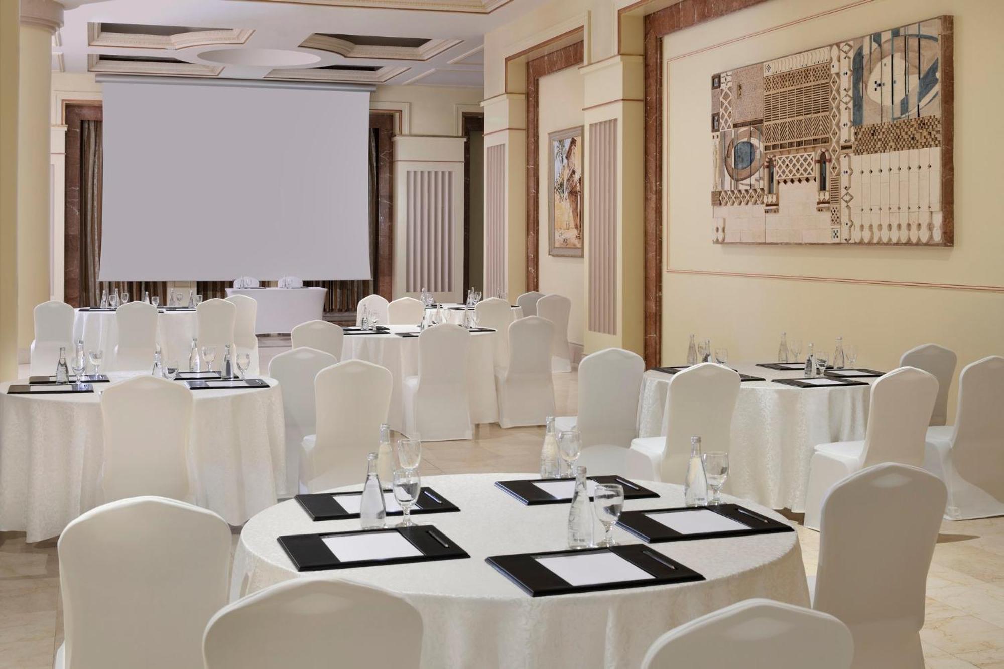 Intercontinental Dar Al Iman Madinah By Ihg Hotel Medina Exterior foto The photo shows a well-decorated event space or meeting room. It features several round tables covered with white tablecloths and surrounded by white chairs. Each table has black placemats and glass water bottles, suggesting a formal setup for a gath