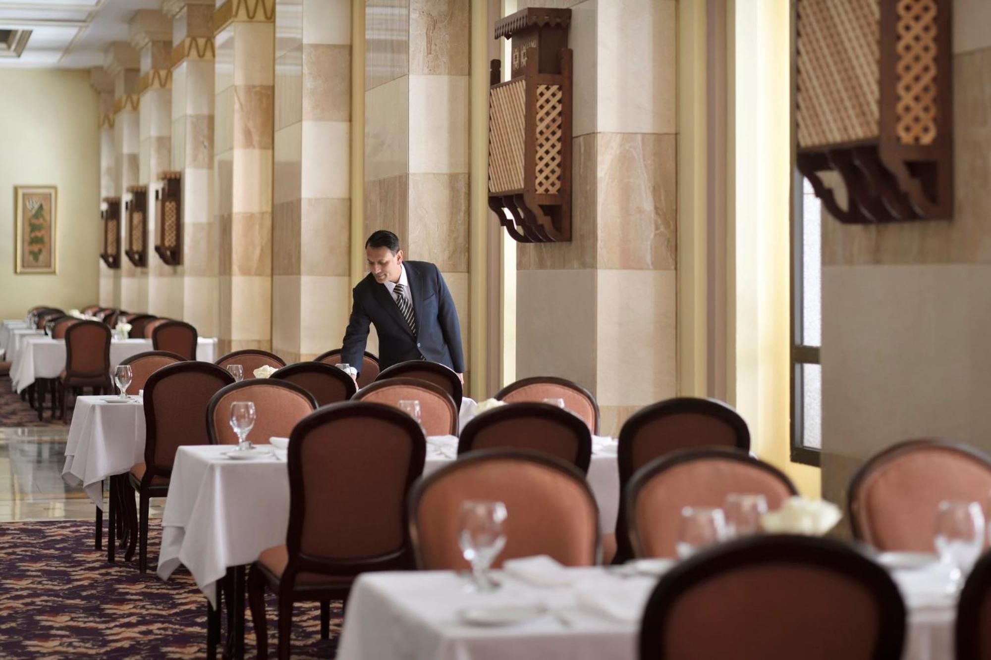 Intercontinental Dar Al Iman Madinah By Ihg Hotel Medina Exterior foto The photo depicts a dining area in an elegant restaurant. There are several tables set with white tablecloths, each accompanied by chairs. A waiter, dressed in a formal suit, is seen arranging or tending to one of the tables. The restaurant features 