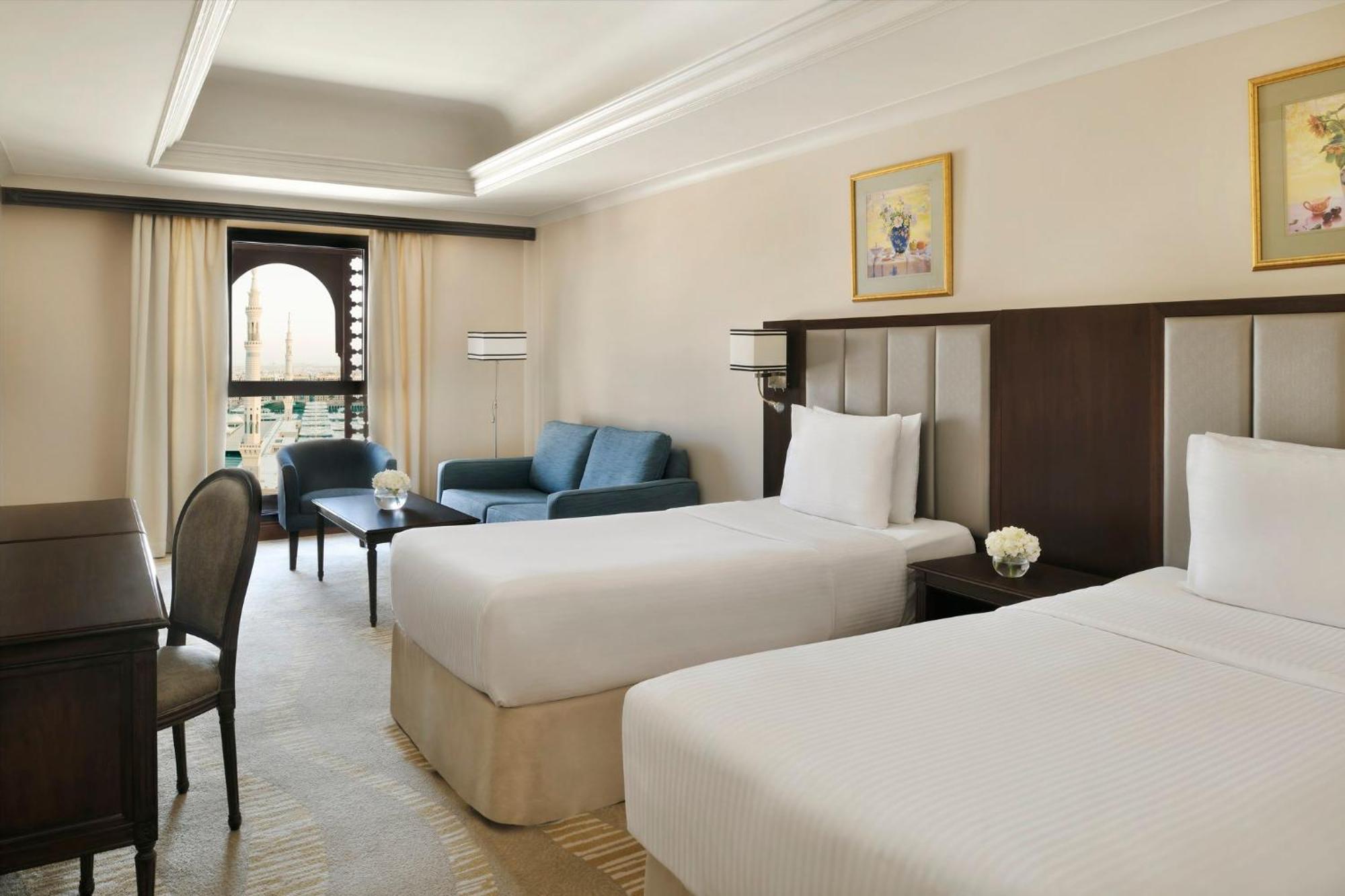 Intercontinental Dar Al Iman Madinah By Ihg Hotel Medina Exterior foto The photo shows a well-appointed hotel room featuring two twin beds with white bedding. There is a small desk with a chair, a comfortable blue sofa, and a coffee table in the sitting area. The room has a warm, neutral color scheme, accented by two pa