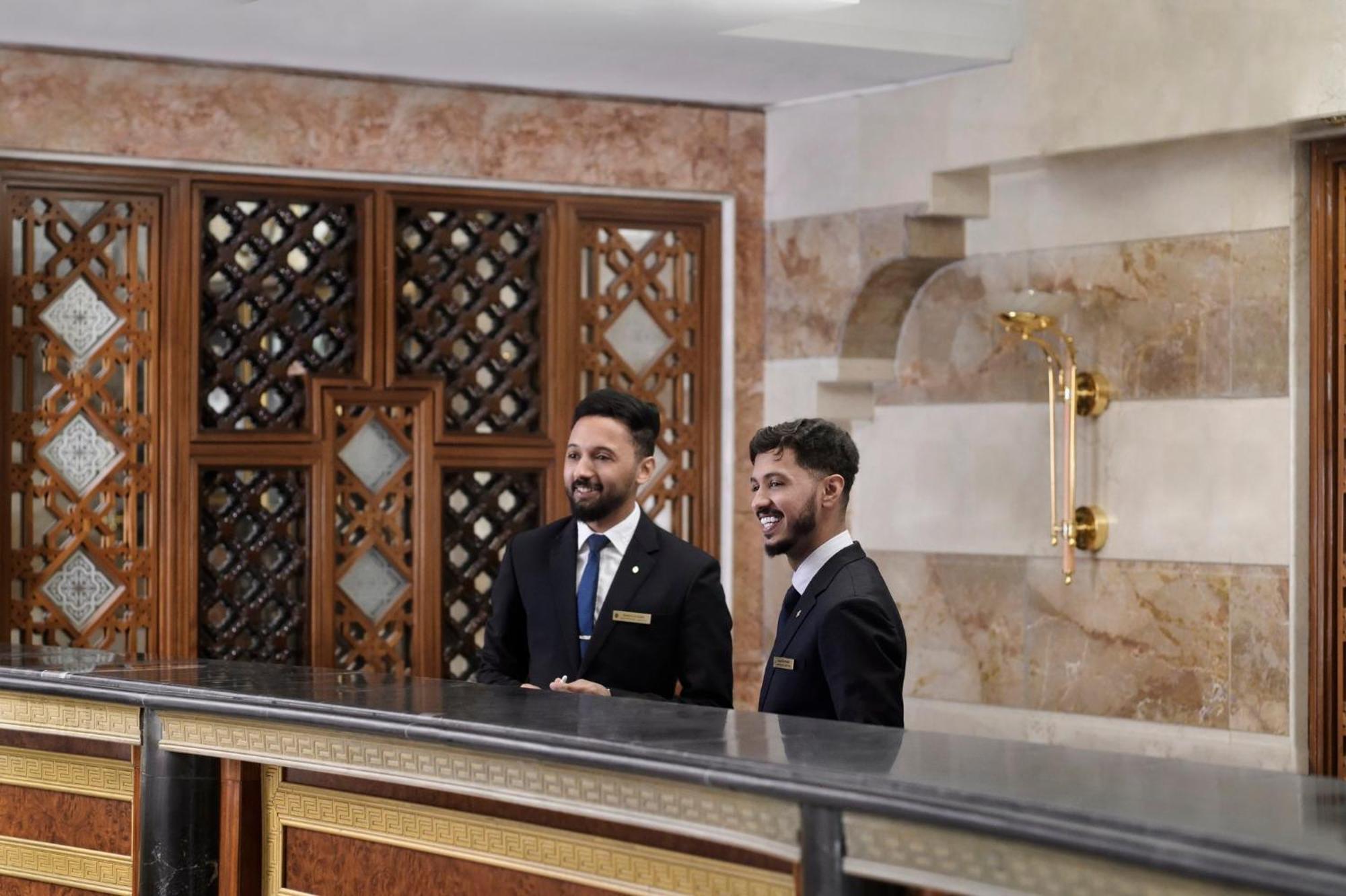 Intercontinental Dar Al Iman Madinah By Ihg Hotel Medina Exterior foto The photo shows two men standing behind a reception desk in an elegant setting, likely a hotel or similar establishment. They are both dressed in formal attire, wearing suits and ties, and appear to be smiling or engaging in conversation. The backgro