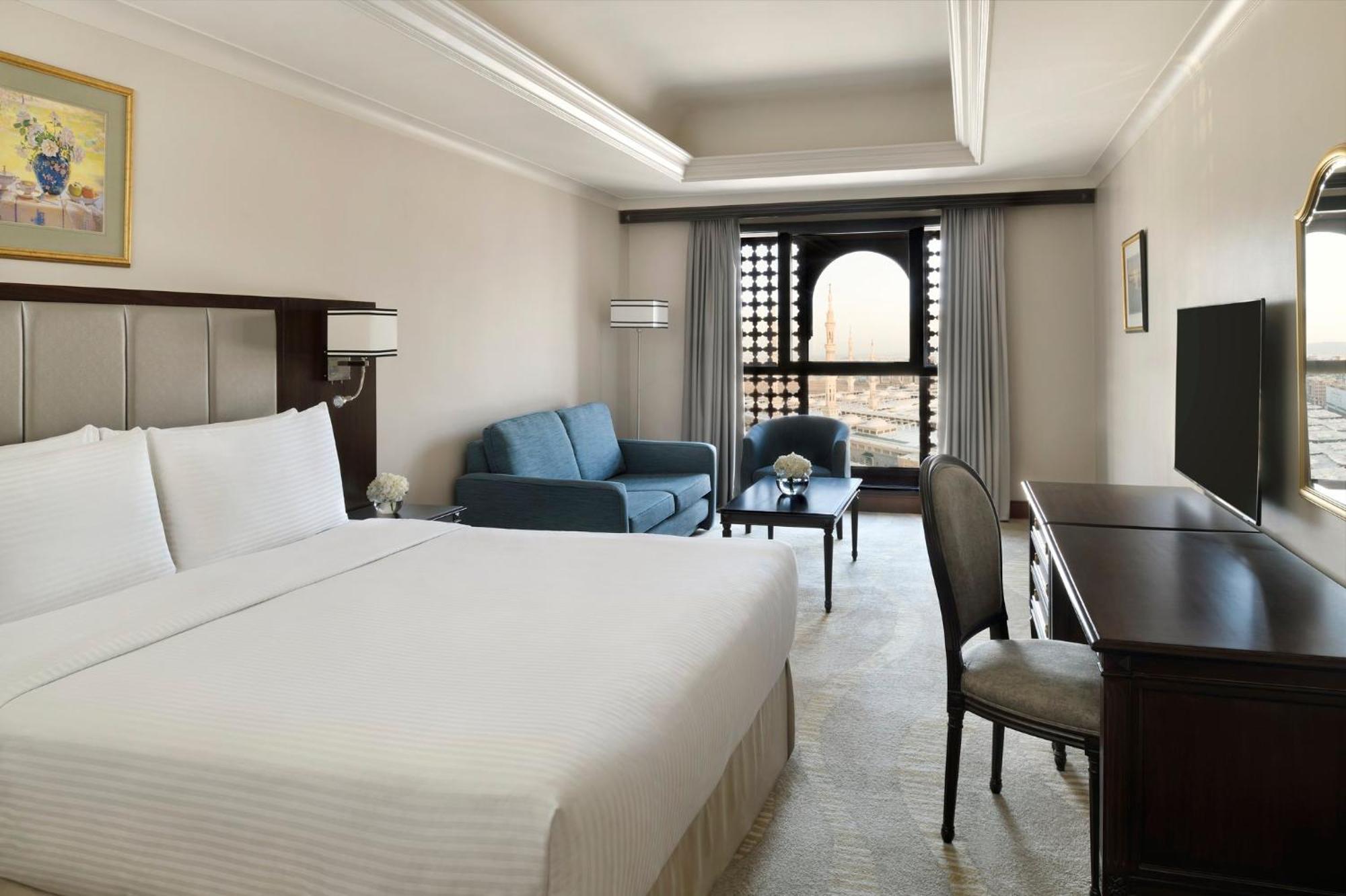 Intercontinental Dar Al Iman Madinah By Ihg Hotel Medina Exterior foto The photo shows a hotel room designed with a modern and comfortable aesthetic. It features a large bed with white linens, a seating area with a blue sofa, and a small table. There is a wooden desk with a chair and a television opposite the bed. Natur