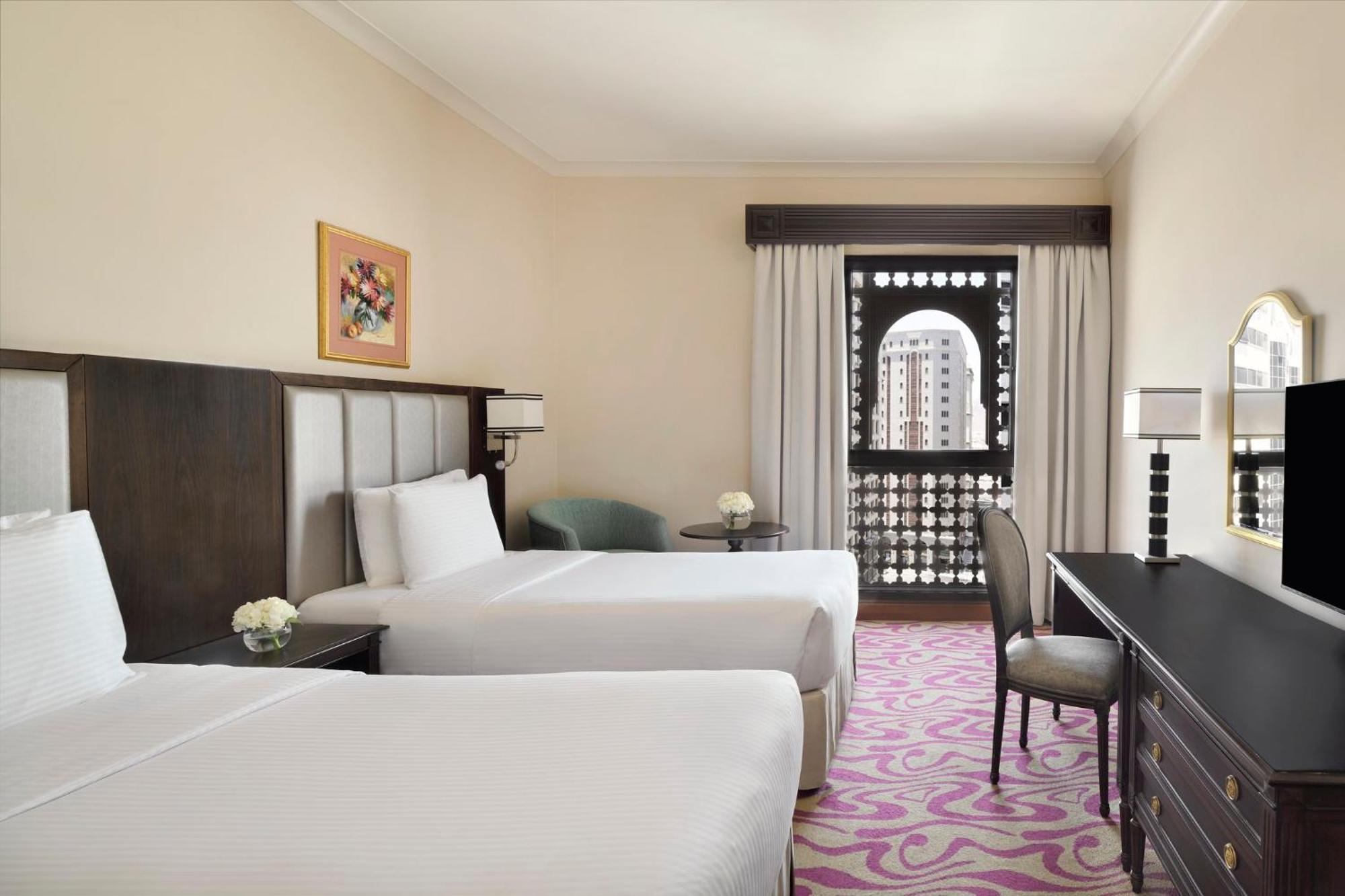 Intercontinental Dar Al Iman Madinah By Ihg Hotel Medina Exterior foto The photo shows a hotel room featuring two twin beds with white bedding. There is a decorative piece of art on the wall above one of the beds. A small round table with a flower arrangement is placed next to the beds. A desk and chair are situated aga