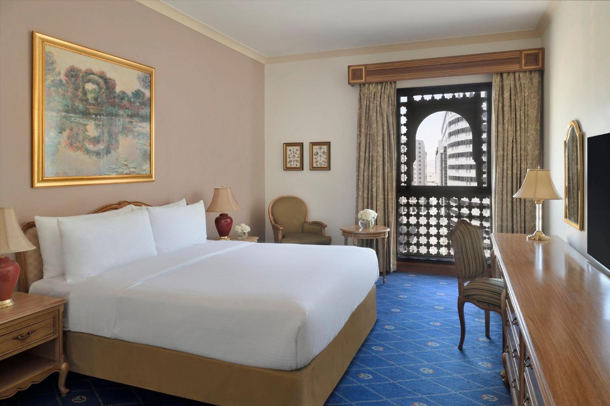 Intercontinental Dar Al Iman Madinah By Ihg Hotel Medina Exterior foto The photo shows a hotel room that features a large, neatly made bed with white linens. There is a framed painting on the wall above the bed. To the side, a small seating area includes a chair and a side table. Opposite the bed, there is a wooden desk