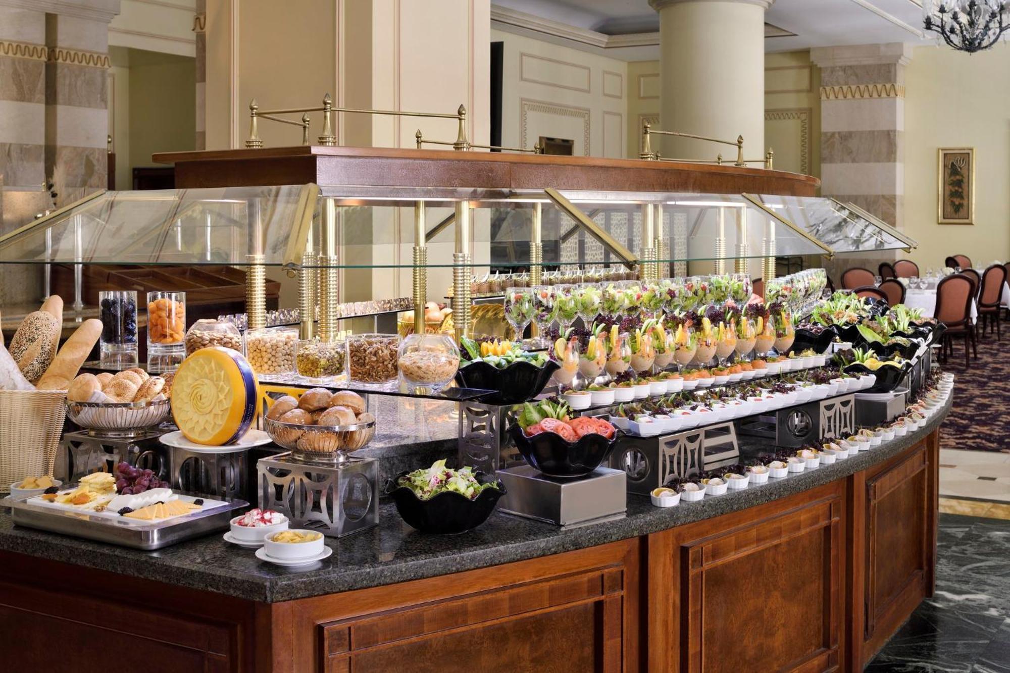 Intercontinental Dar Al Iman Madinah By Ihg Hotel Medina Exterior foto The photo shows an elegantly arranged buffet with a variety of food items. There is a large, curved wooden display with glass covers, showcasing an assortment of dishes. These include different types of breads and rolls, salads, garnishes, and desser