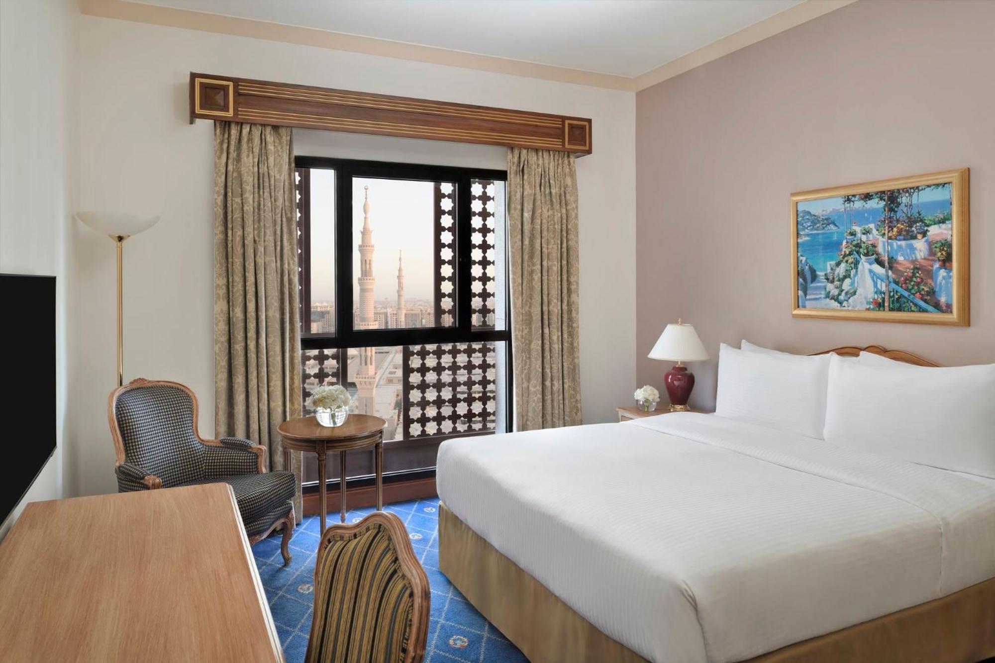 Intercontinental Dar Al Iman Madinah By Ihg Hotel Medina Exterior foto The photo shows a hotel room with a modern design. There is a large bed with white linens placed against the wall. Next to the bed, there is a small bedside table with a lamp. A comfortable armchair is situated nearby, accompanied by a wooden desk. T