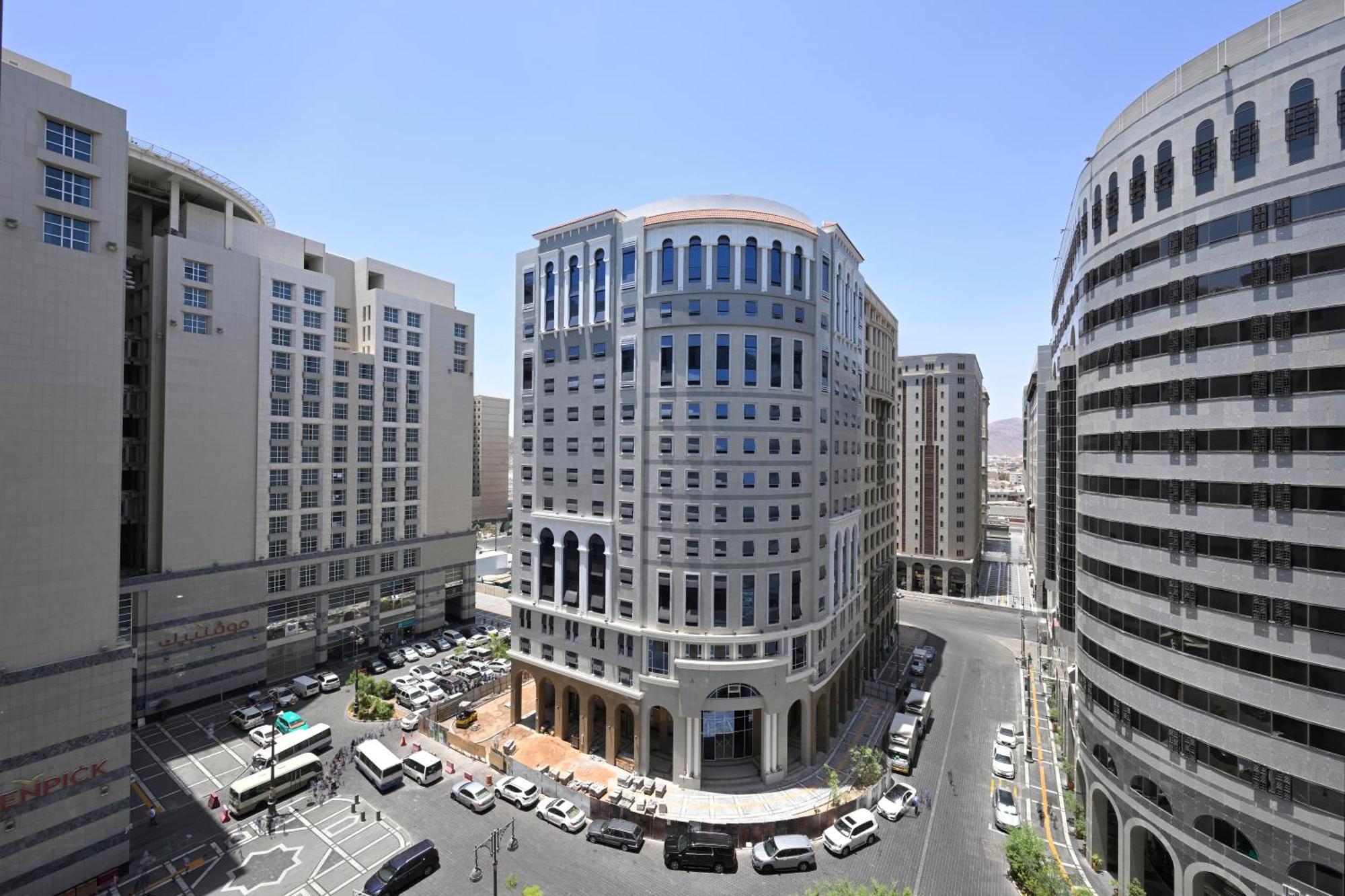 Intercontinental Dar Al Iman Madinah By Ihg Hotel Medina Exterior foto The photo shows a modern urban scene featuring several buildings, including a prominent rounded building with a glass façade. This building appears to be situated at an intersection, surrounded by other high-rise structures that have distinct archite