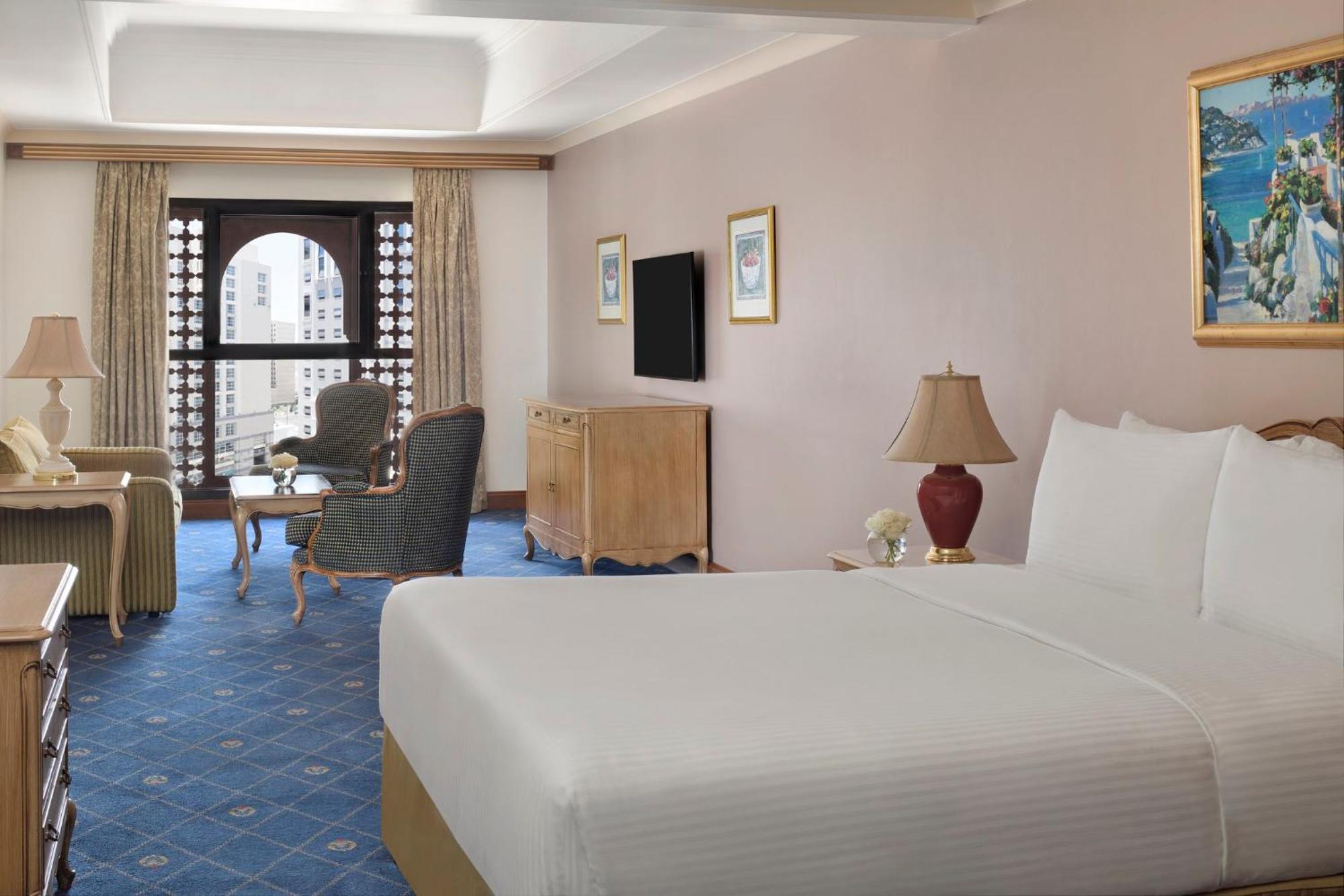 Intercontinental Dar Al Iman Madinah By Ihg Hotel Medina Exterior foto The photo shows a well-furnished hotel room. It features a large bed with a white duvet, two bedside lamps, and decorative paintings on the walls. There is a comfortable seating area with two chairs and a small table. A wooden dresser is positioned a