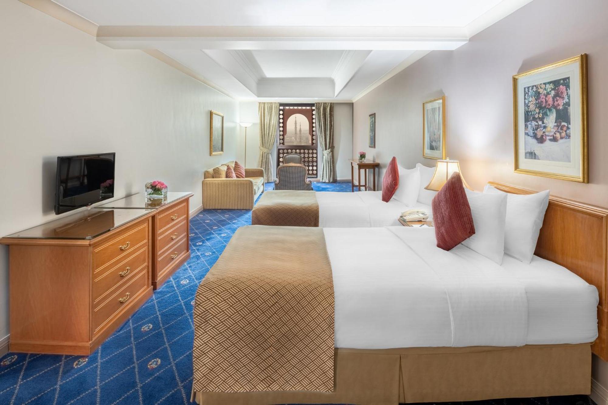 Intercontinental Dar Al Iman Madinah By Ihg Hotel Medina Exterior foto The photo shows a hotel room featuring two beds with white linens and decorative pillows. There is a wooden dresser with a television on it. The flooring has a blue carpet with a pattern, and the walls are painted in a light color. The room is well-l