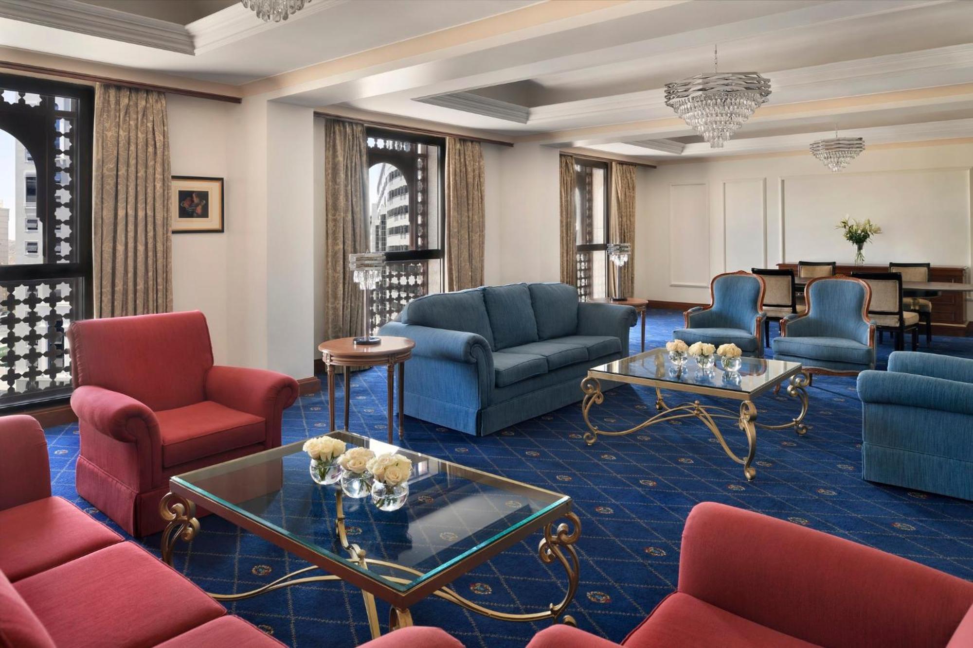 Intercontinental Dar Al Iman Madinah By Ihg Hotel Medina Exterior foto The photo shows a luxurious living space or lounge area. It features a combination of furniture, including a blue sofa, several red armchairs, and glass-top coffee tables with gold accents. The flooring is carpeted in a deep blue, and the walls are a