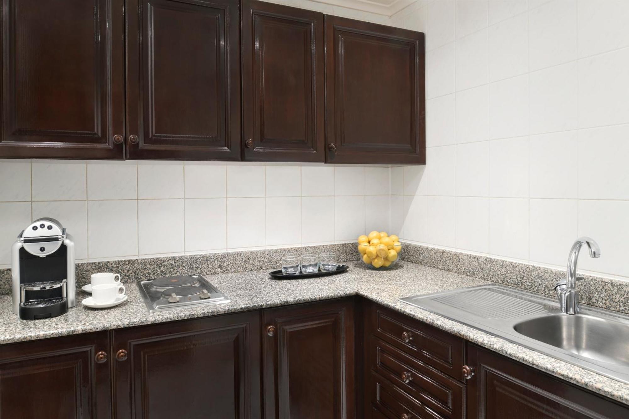 Intercontinental Dar Al Iman Madinah By Ihg Hotel Medina Exterior foto The photo shows a kitchen with dark wooden cabinetry and a light-colored countertop. There are a few appliances visible, including a coffee machine and a stovetop with burners. To the right, there is a sink, and a bowl of yellow lemons is placed on t