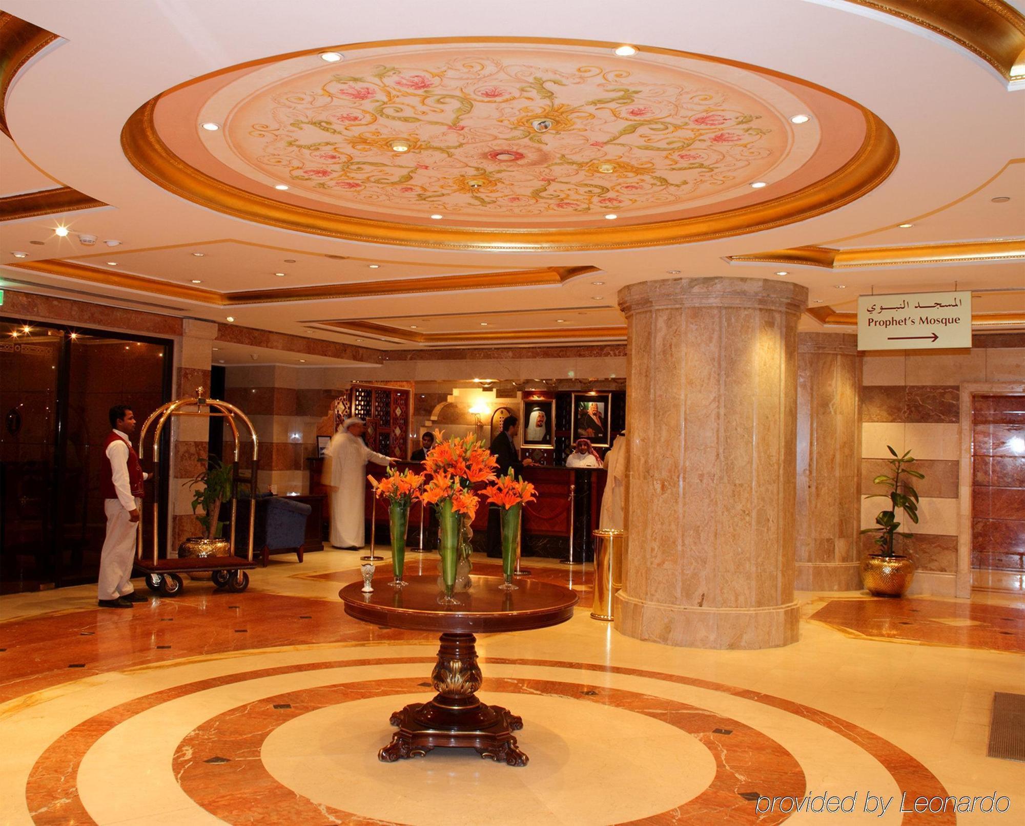 Intercontinental Dar Al Iman Madinah By Ihg Hotel Medina Interior foto The photo depicts a luxurious hotel lobby. The ceiling features an ornate design with floral patterns, adding elegance to the space. In the center of the lobby, there is a decorative table with a bowl or vase of bright orange flowers. The flooring is