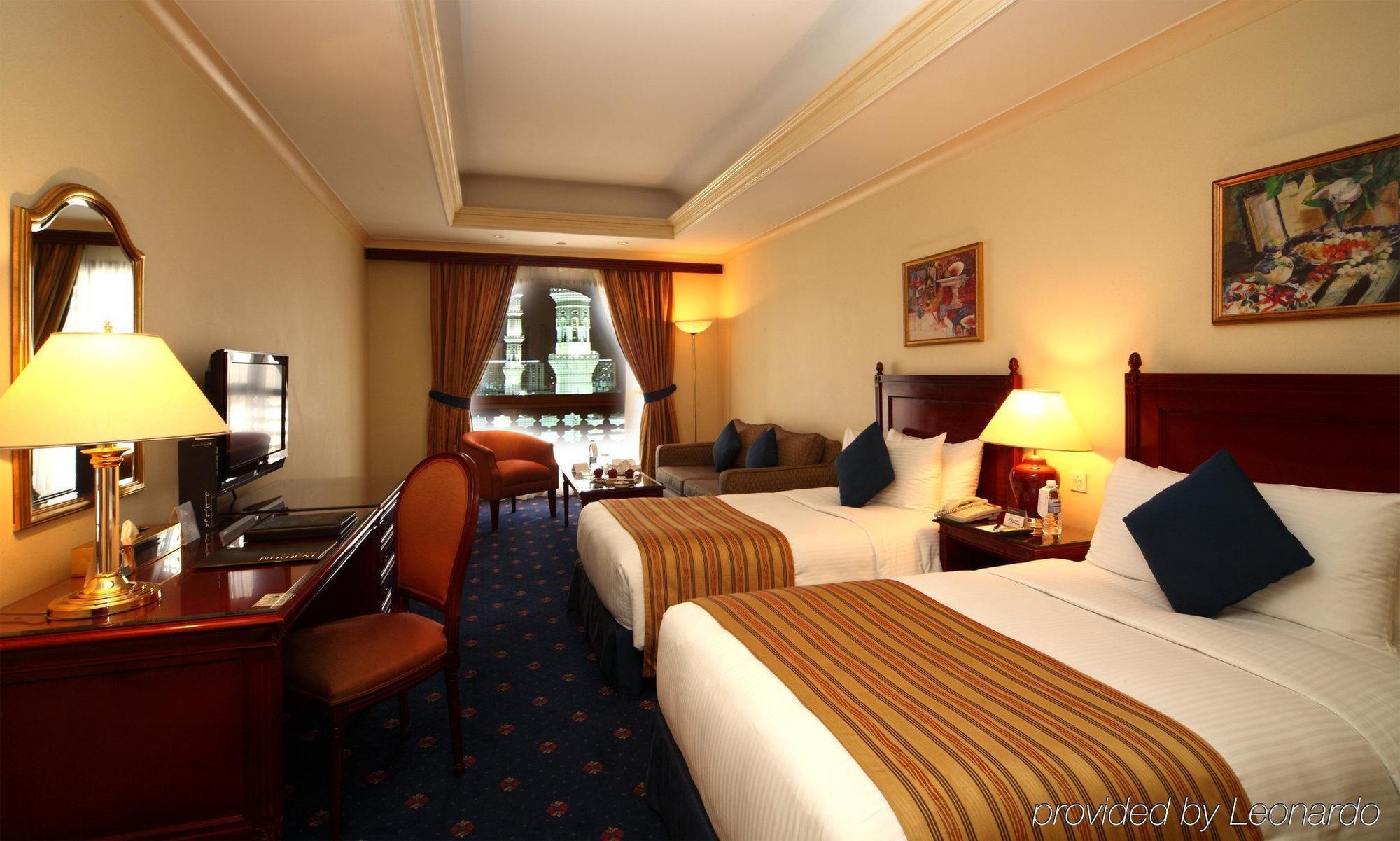 Intercontinental Dar Al Iman Madinah By Ihg Hotel Medina Cameră foto The photo shows a hotel room featuring two beds with white linens and brown striped throws. There is a wooden desk and chair, alongside a television. The walls are painted in a warm tone, and there are decorative paintings. Large windows with curtain