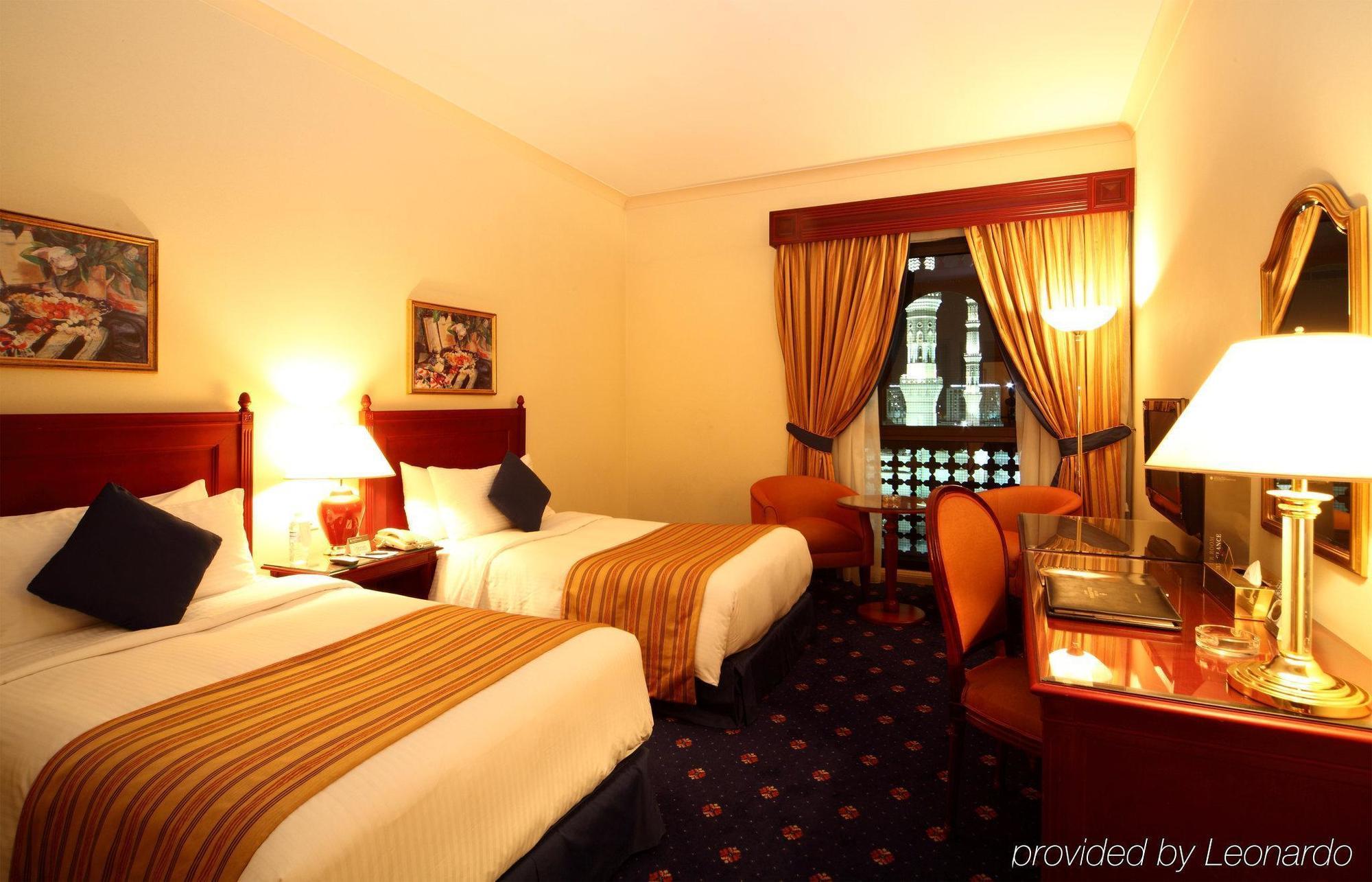 Intercontinental Dar Al Iman Madinah By Ihg Hotel Medina Cameră foto The photo shows a hotel room featuring two twin beds with white linens and brown striped bedspreads. There are decorative pillows on the beds, and the room has warm-colored walls. To the side, there's a small sitting area with a round table and a cha