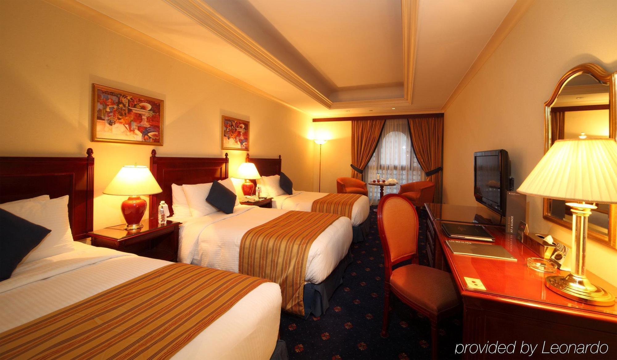 Intercontinental Dar Al Iman Madinah By Ihg Hotel Medina Cameră foto The photo shows a hotel room with a warm and inviting atmosphere. There are two beds with white and gold bedding, complemented by decorative pillows. Each bed has a bedside lamp. In the background, there's a small seating area with a couple of chairs