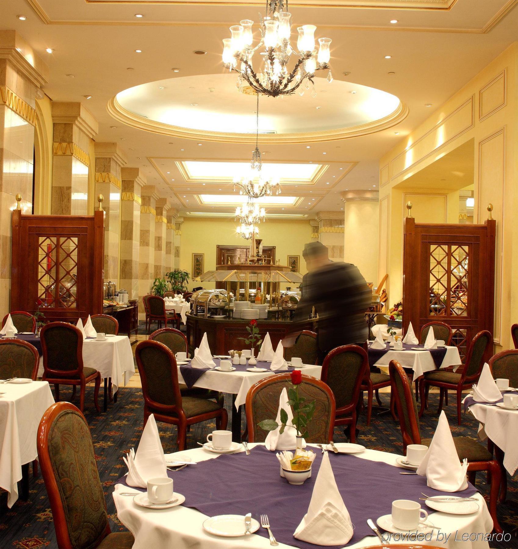 Intercontinental Dar Al Iman Madinah By Ihg Hotel Medina Restaurant foto The photo depicts an elegant restaurant interior. It features round tables covered with white tablecloths, each set with plates, cutlery, and folded napkins arranged into triangles. The space has a warm, inviting atmosphere with a mix of decorative e