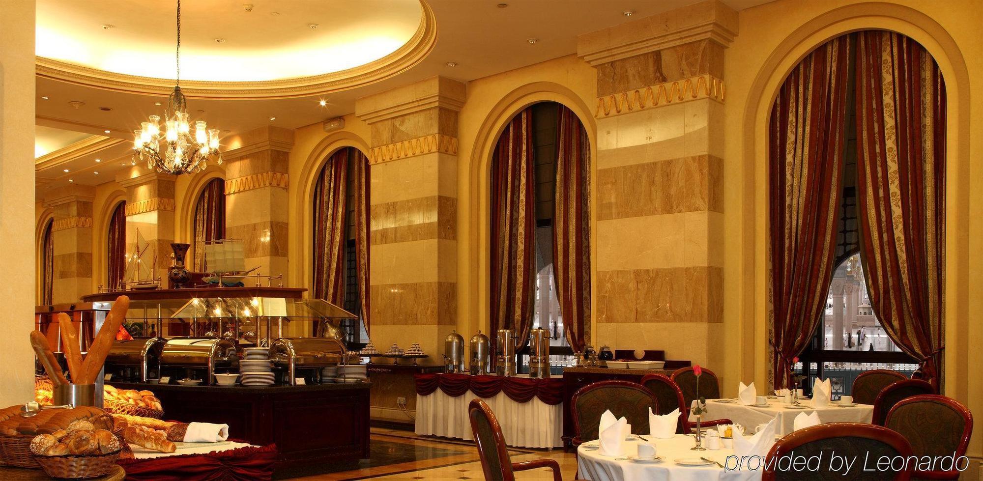 Intercontinental Dar Al Iman Madinah By Ihg Hotel Medina Restaurant foto The photo shows an elegant dining restaurant interior. It features stylish arches and walls with a blend of marble and decorative elements. There are large windows draped with rich curtains, contributing to the luxurious atmosphere. The dining area i