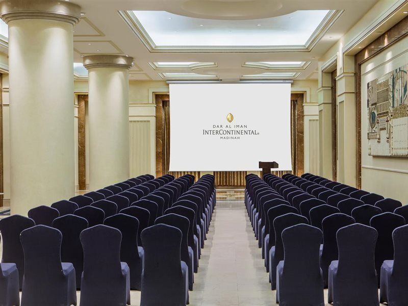 Intercontinental Dar Al Iman Madinah By Ihg Hotel Medina Exterior foto The photo shows a conference or meeting room set up. There are rows of dark blue chairs arranged facing a large screen at the front of the room. The screen displays the logo of the Intercontinental hotel, indicating that this space is likely intended