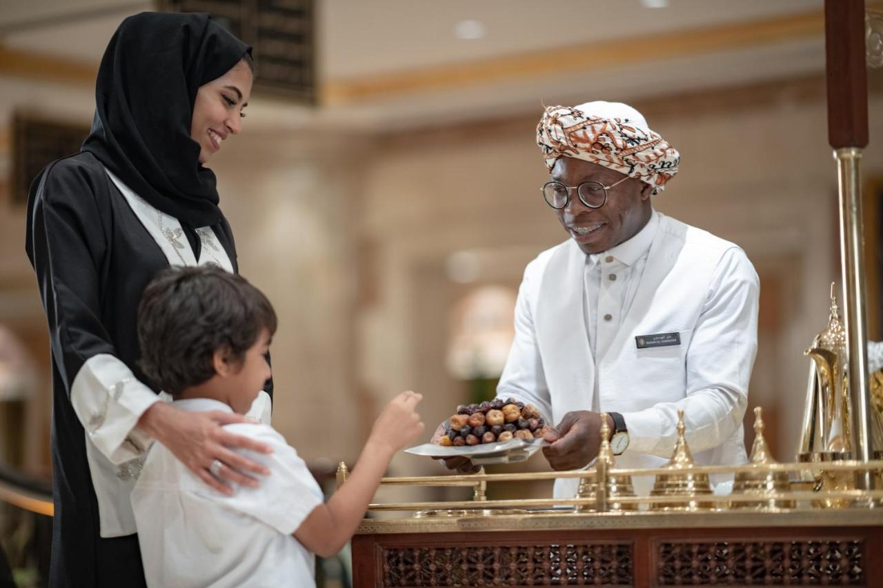 Intercontinental Dar Al Iman Madinah By Ihg Hotel Medina Exterior foto The photo depicts a warm interaction between three people. A woman, dressed in a traditional black attire and a headscarf, stands beside a young boy who is reaching out towards a plate of dates held by a smiling man in a white traditional outfit with